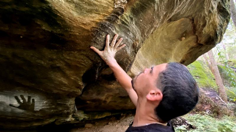 A young adult man stands beneath a rock overhang and touches the rock with his hand. On the rock is another hand print. In the background are more rocks and trees.